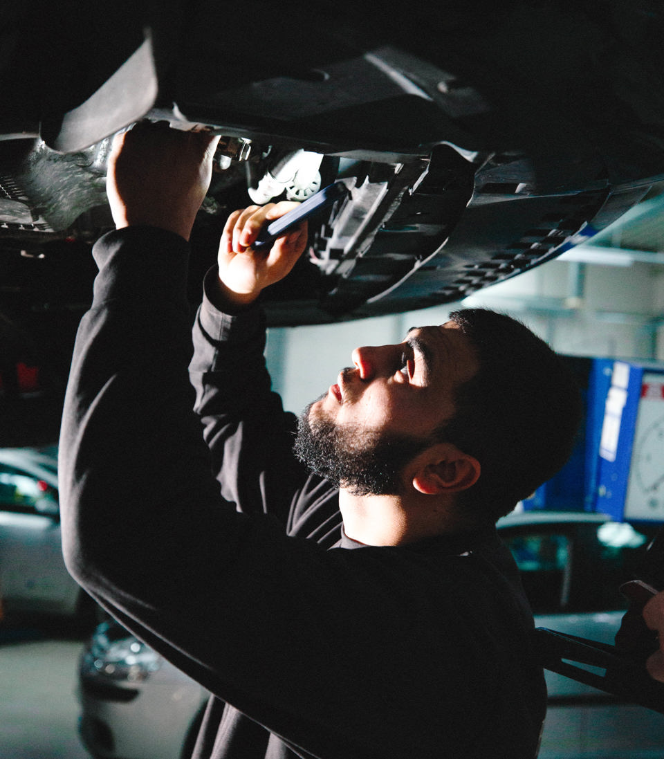 Lærling arbejder under en bil, der er hævet på en løfteplatform i værkstedet, Lærling, bil, løfteplatform, værksted, bilreparation, mekanikeruddannelse, personvognsmekaniker, erhvervsskole.