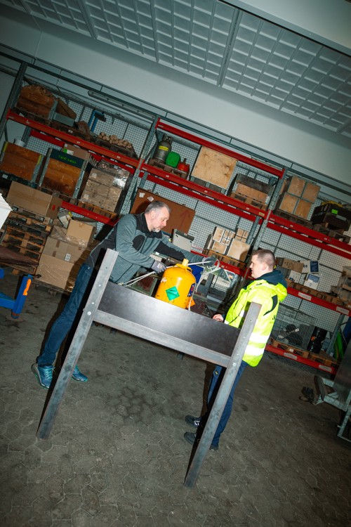 Personer arbejder ved et bord i et lagerområde, Lufthavnsuddannelsen,  Lagerarbejde, sikkerhedsvest, teamwork, arbejdsstation, lufthavn, logistik, opbevaring, samarbejde