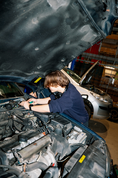 En studerende på GF1-uddannelsen på en erhvervsskole arbejder koncentreret under motorhjelmen på en bil i et værksted, Motorhjelm, Værksted, Bilmekanik, Automekaniker, Hands-on læring,  Bilreparation,