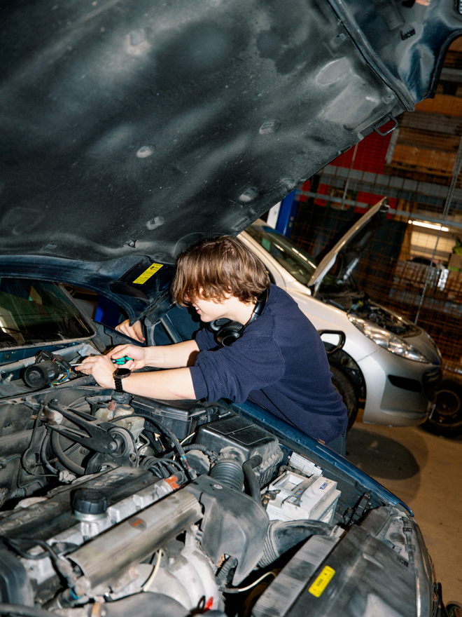 En studerende på GF1-uddannelsen på en erhvervsskole arbejder koncentreret under motorhjelmen på en bil i et værksted, Motorhjelm, Værksted, Bilmekanik, Automekaniker, Hands-on læring, Bilreparation,
