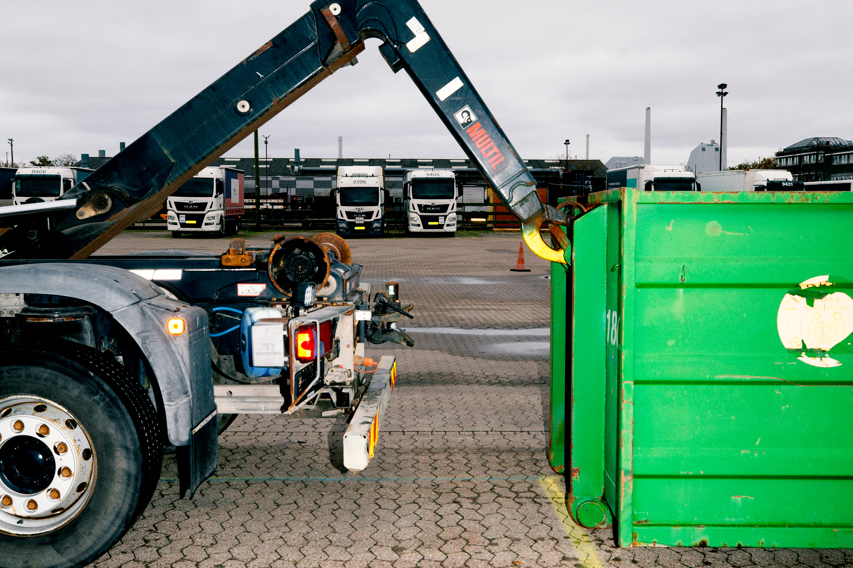 En lastbil med en kranarm er i færd med at påsætte en stor grøn container, chauffør-uddannelsen, chauffør, vejgods, Godshåndtering,  transport,  uddannelsen, vejgodstransport, ContainerLastbil, Kranarm, c